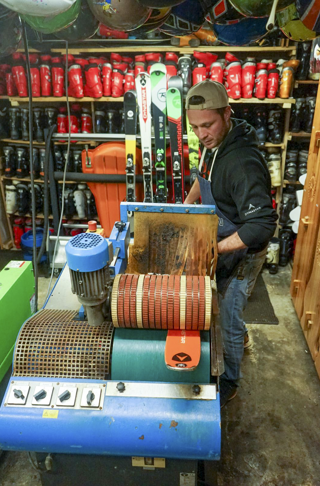Technician in a ski workshop using a maintenance machine to sharpen a ski in front of shelves filled with ski boots.